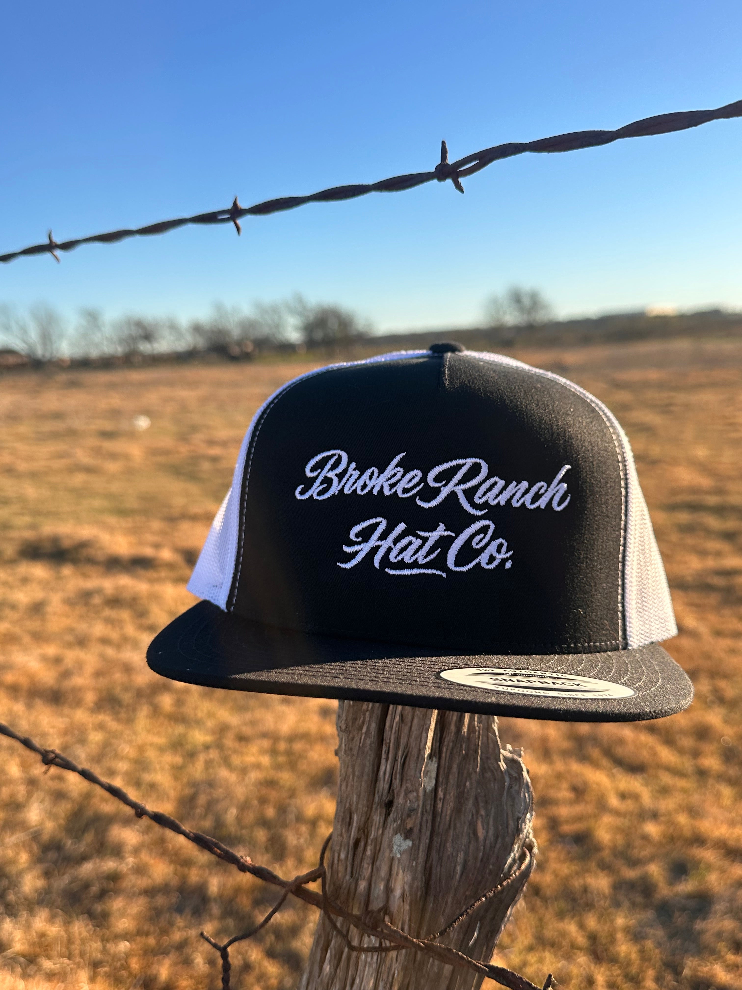 Black and white hat with 'Broke Ranch Hat Co.' text against a barbed wire fence and blue sky background